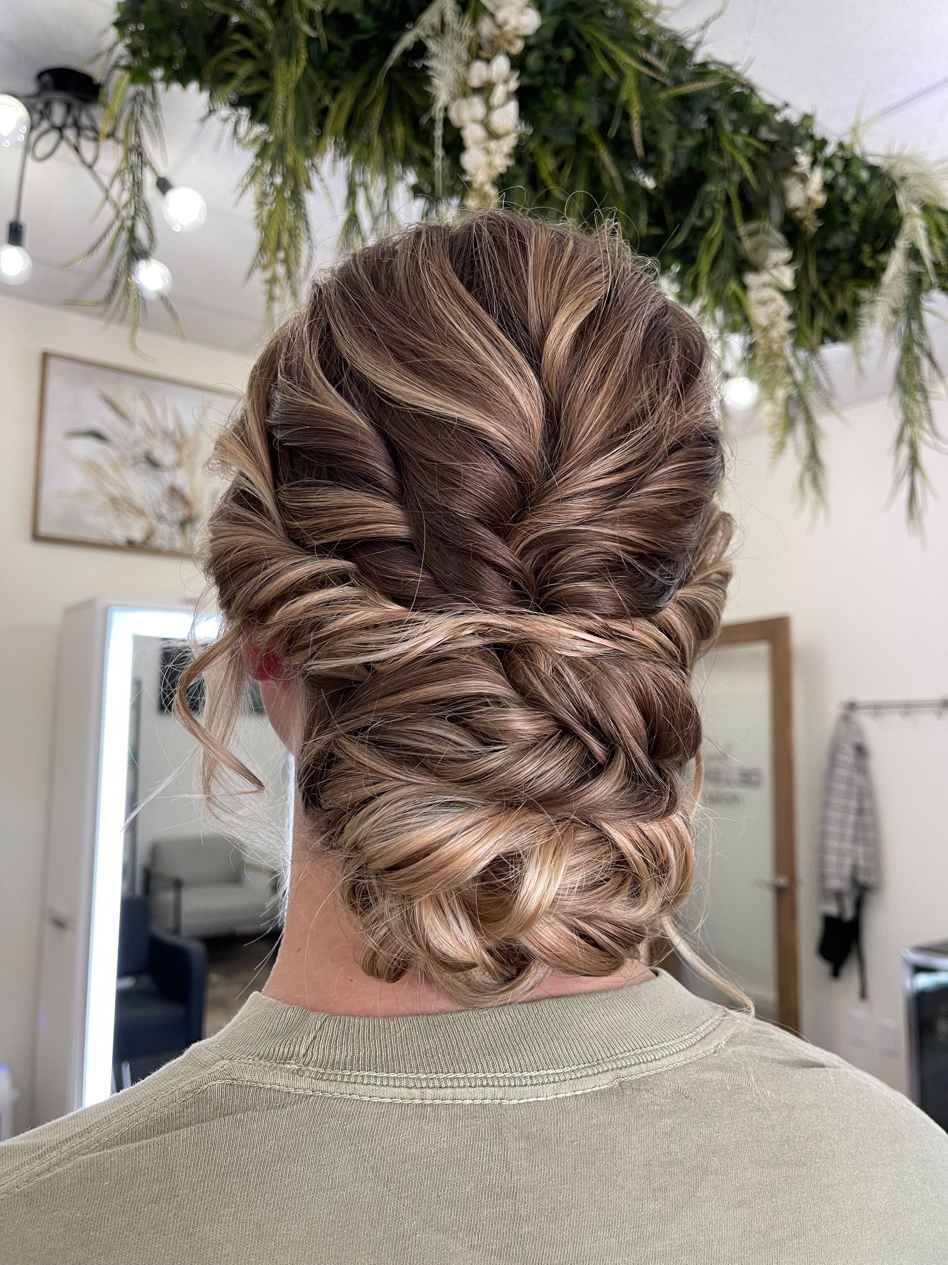 Back view of a person with intricately styled brown and blonde hair in a twisted updo. They are in a salon setting.