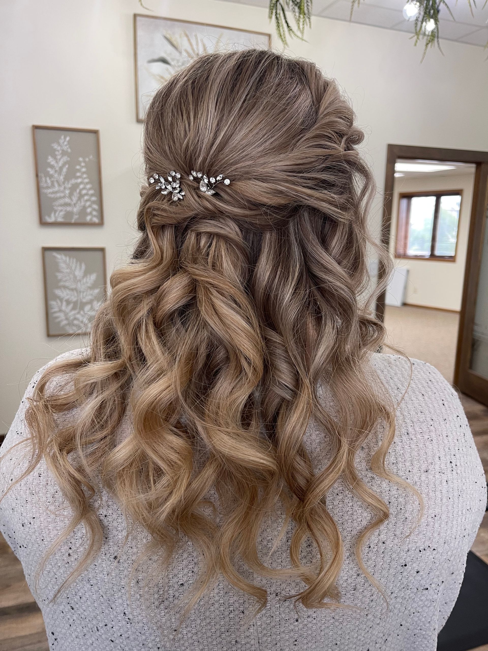 Woman with blonde, curly hair styled half-up, adorned with a small hairpiece.  The woman wears a white, textured top.