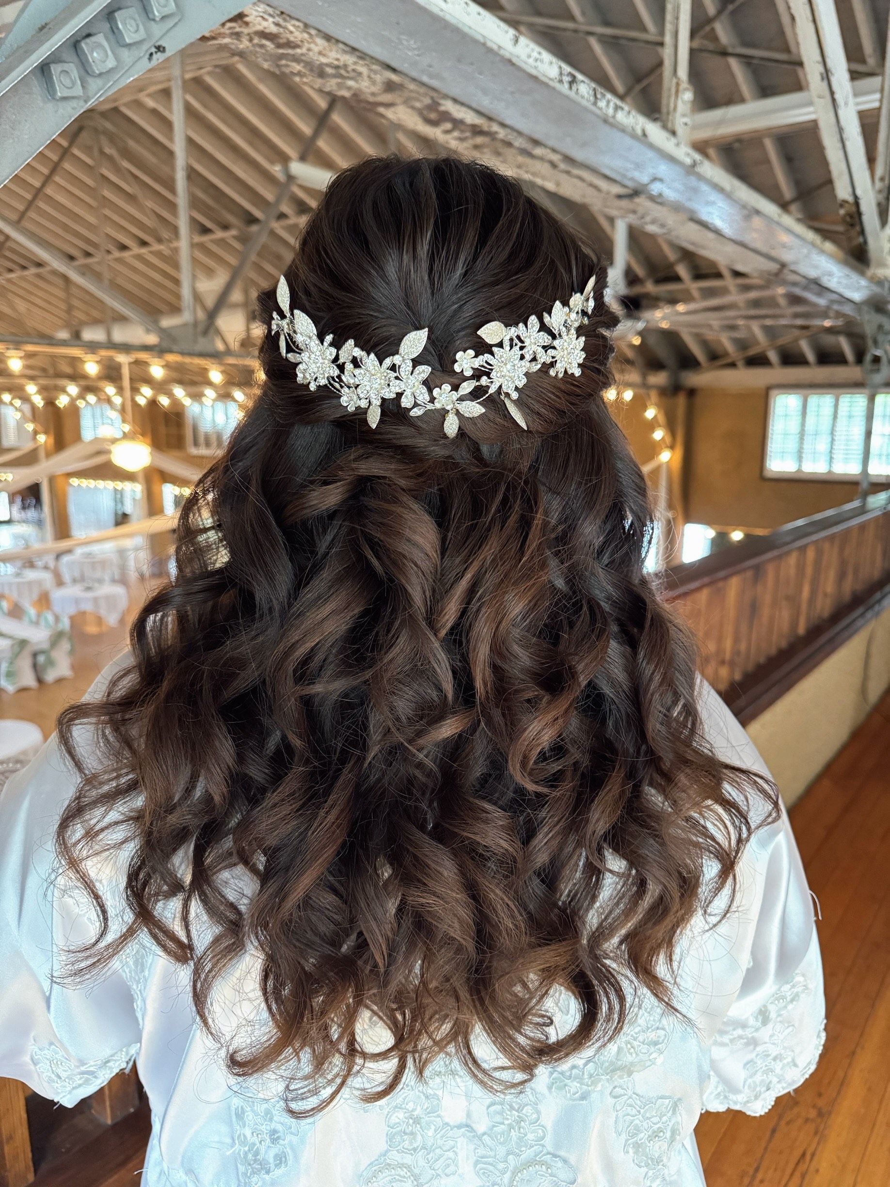 Woman with dark, wavy hair in a half-up style, wearing a delicate floral hairpiece. She’s in a white robe.