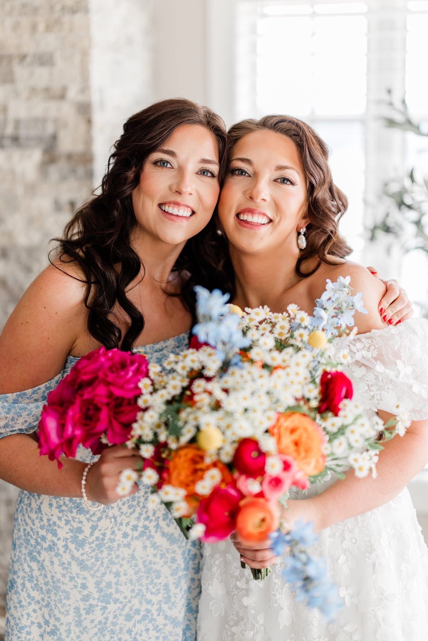 Two smiling women embrace, holding colorful bouquets. One wears a light blue floral dress; the other, a white lace dress.