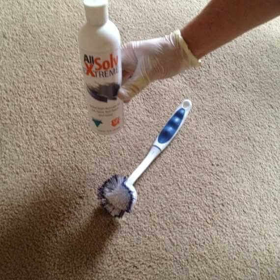 Hand wearing a glove sprays carpet stain remover onto a carpet spot with a scrub brush nearby