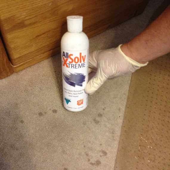 Gloved hand holding a bottle of All Solv Xtreme on a carpeted floor near a wooden surface