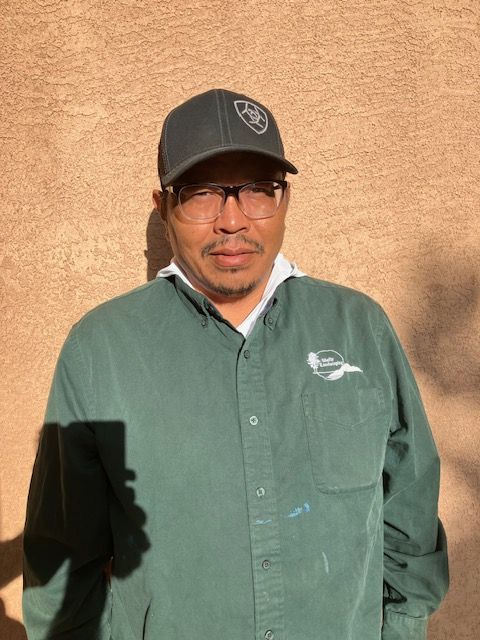 Donovan Gordo - Man wearing glasses and a black cap, standing in sunlight against a tan wall, in a green button-up shirt