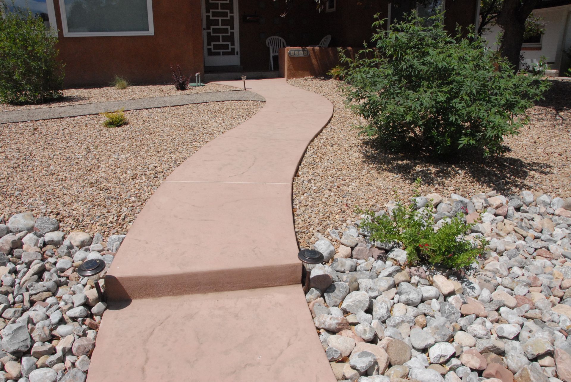 A curved, reddish-brown concrete walkway leading to a house with a step. Gravel and shrubs surround it.