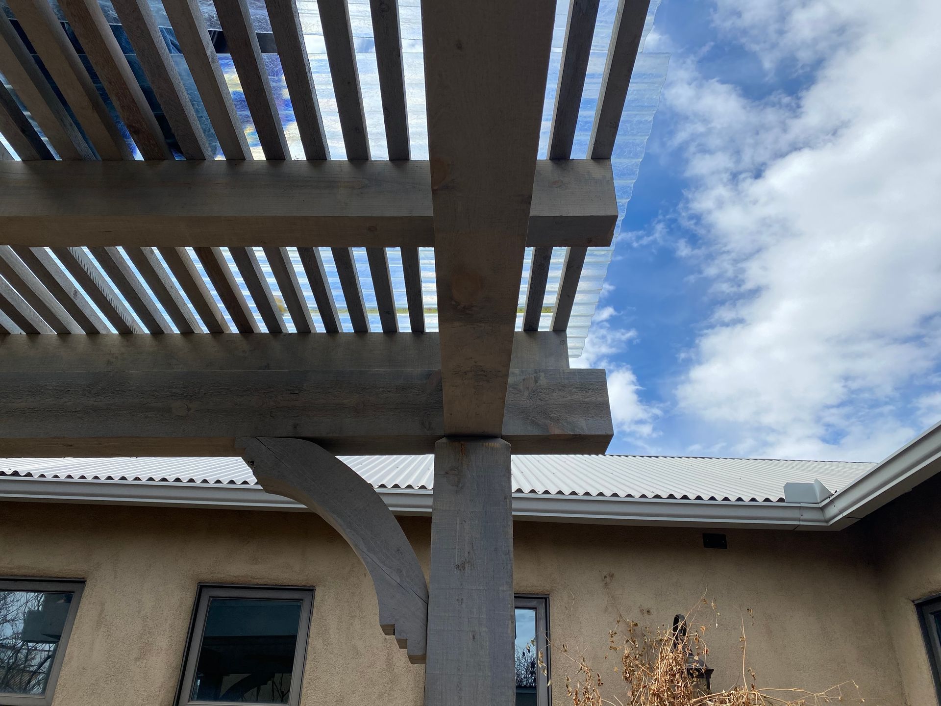 Wooden pergola against a cloudy sky, supported by a post, over a light-colored building.