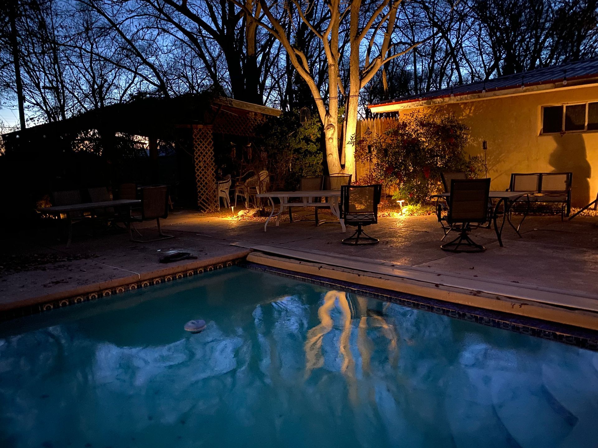 Poolside backyard at dusk, with patio furniture and a lit-up tree reflecting in the water.