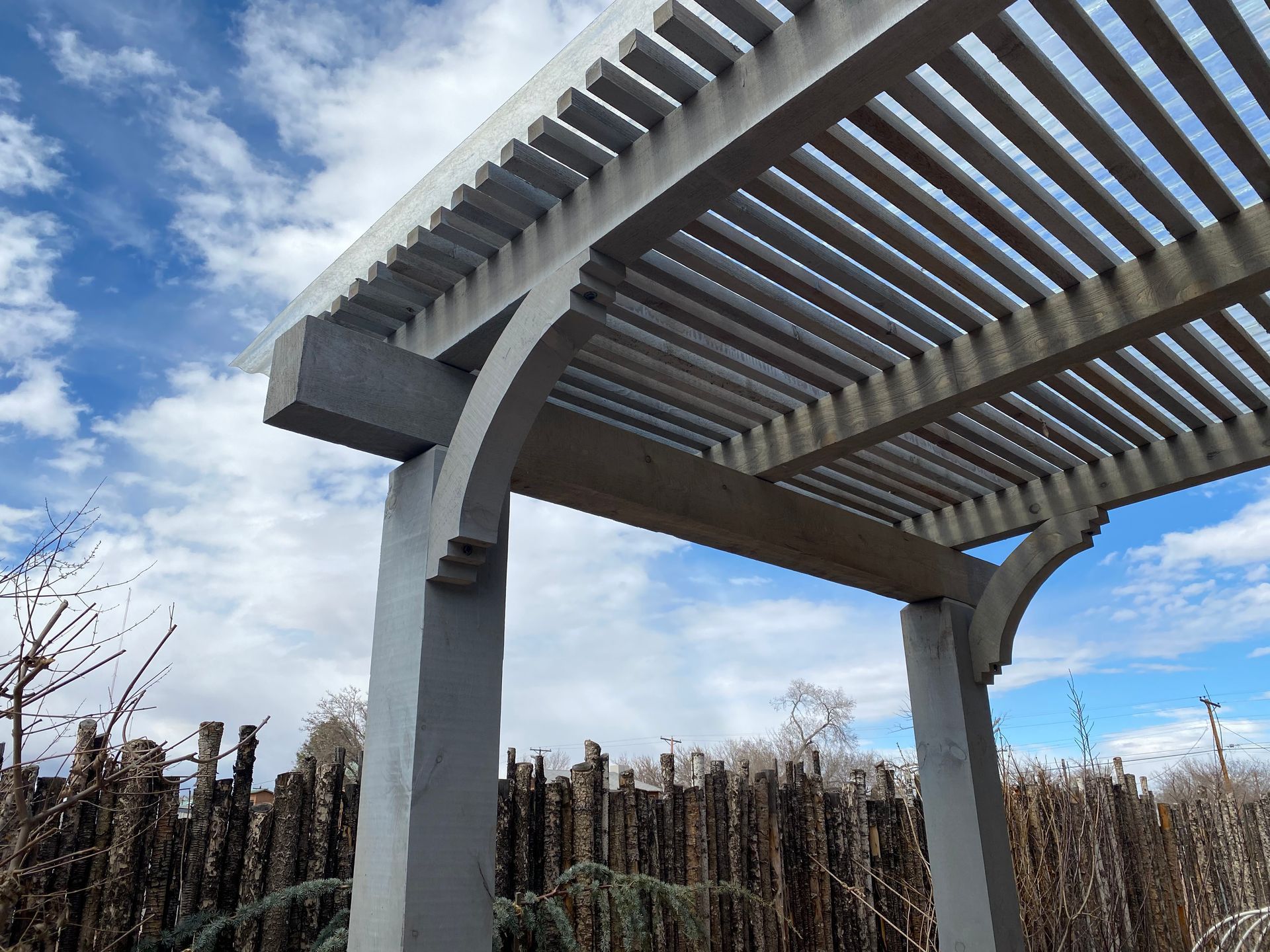 Gray pergola with a slatted roof against a blue sky, in a garden setting.
