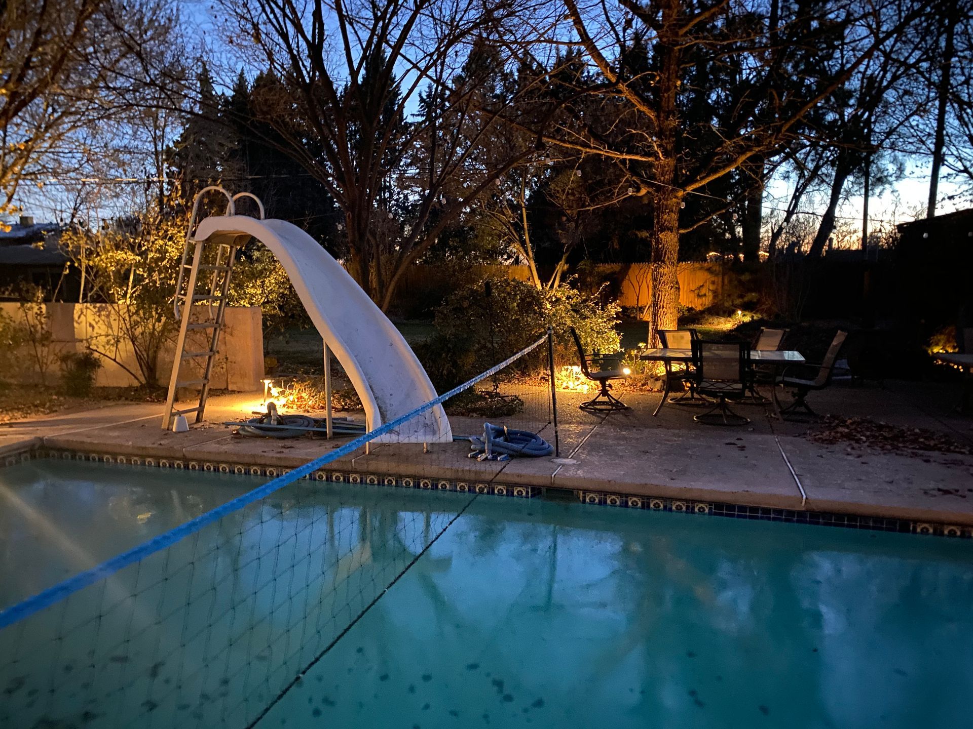 Pool at dusk with slide, lights, and chairs. Blue water, trees, and a dark sky.