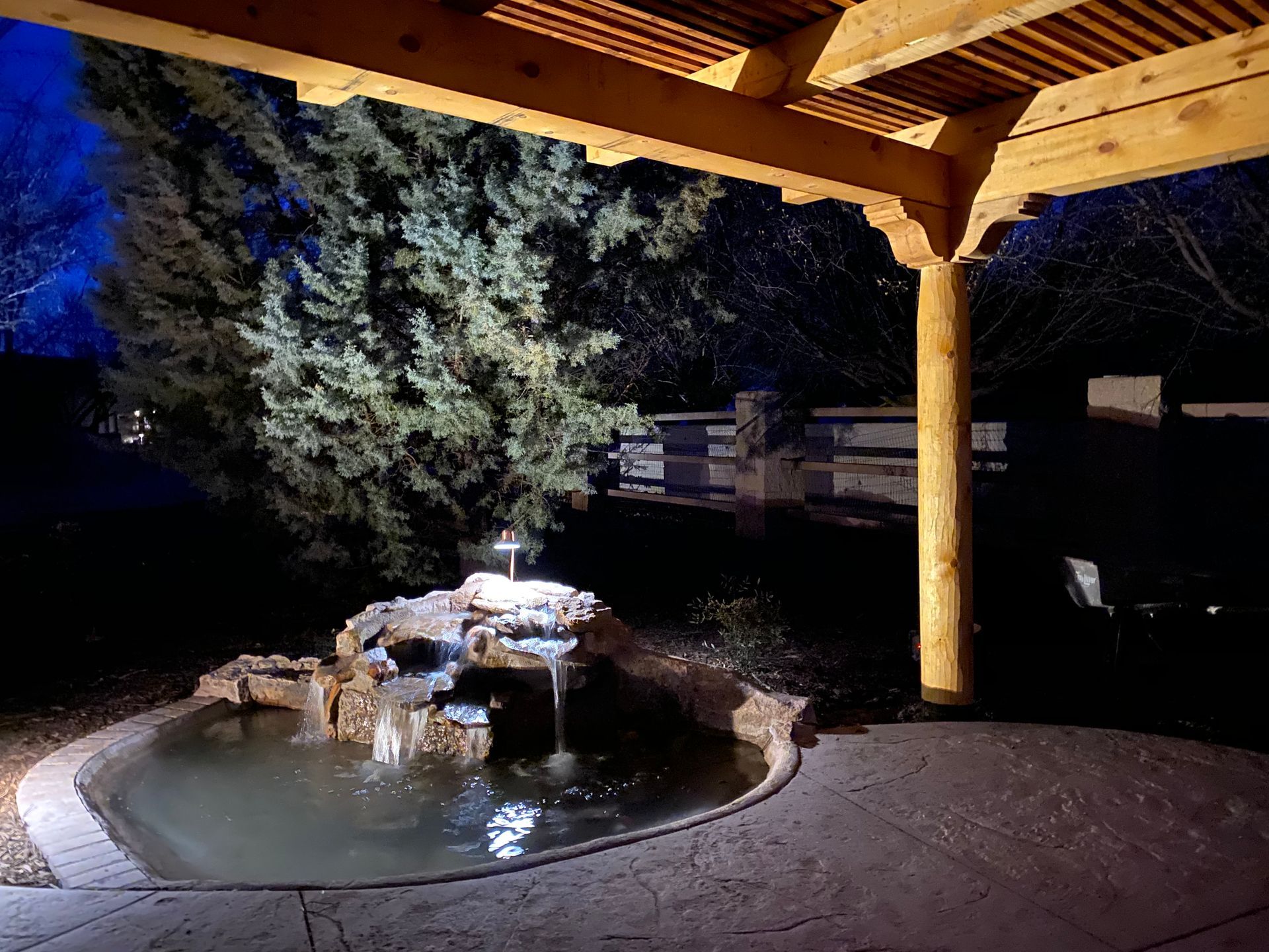Nighttime view of a backyard with a pond, waterfall, and wooden pergola; illuminated.