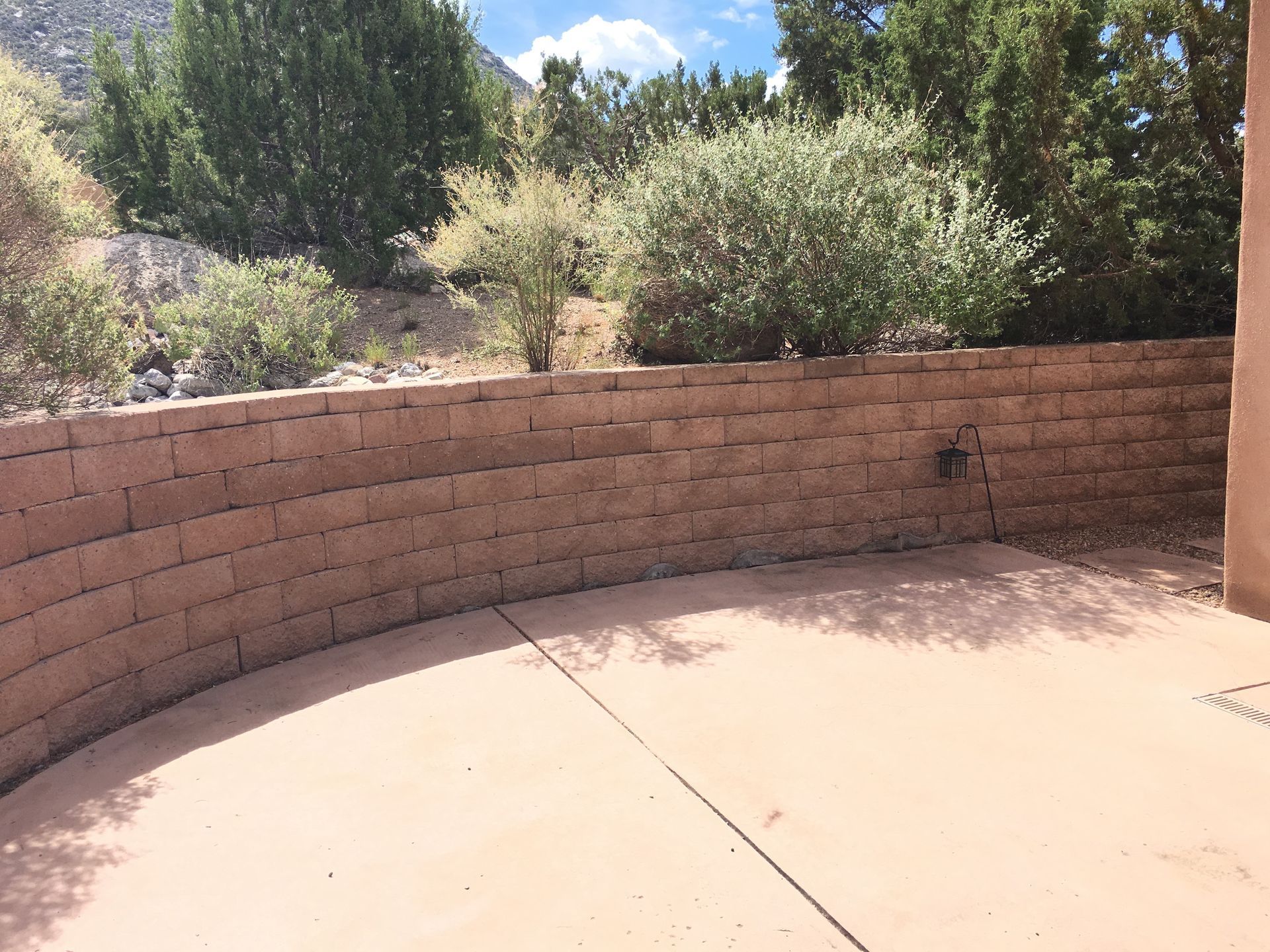 Curved brick retaining wall and concrete patio area with surrounding shrubs and trees under a partly cloudy sky.