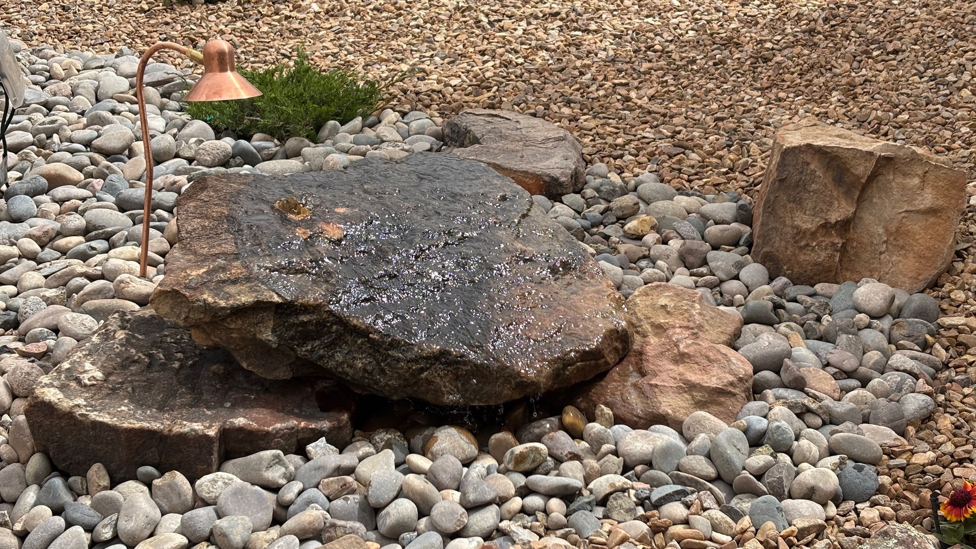 Water feature with dark rocks, pebbles, and copper lamp in an outdoor setting.