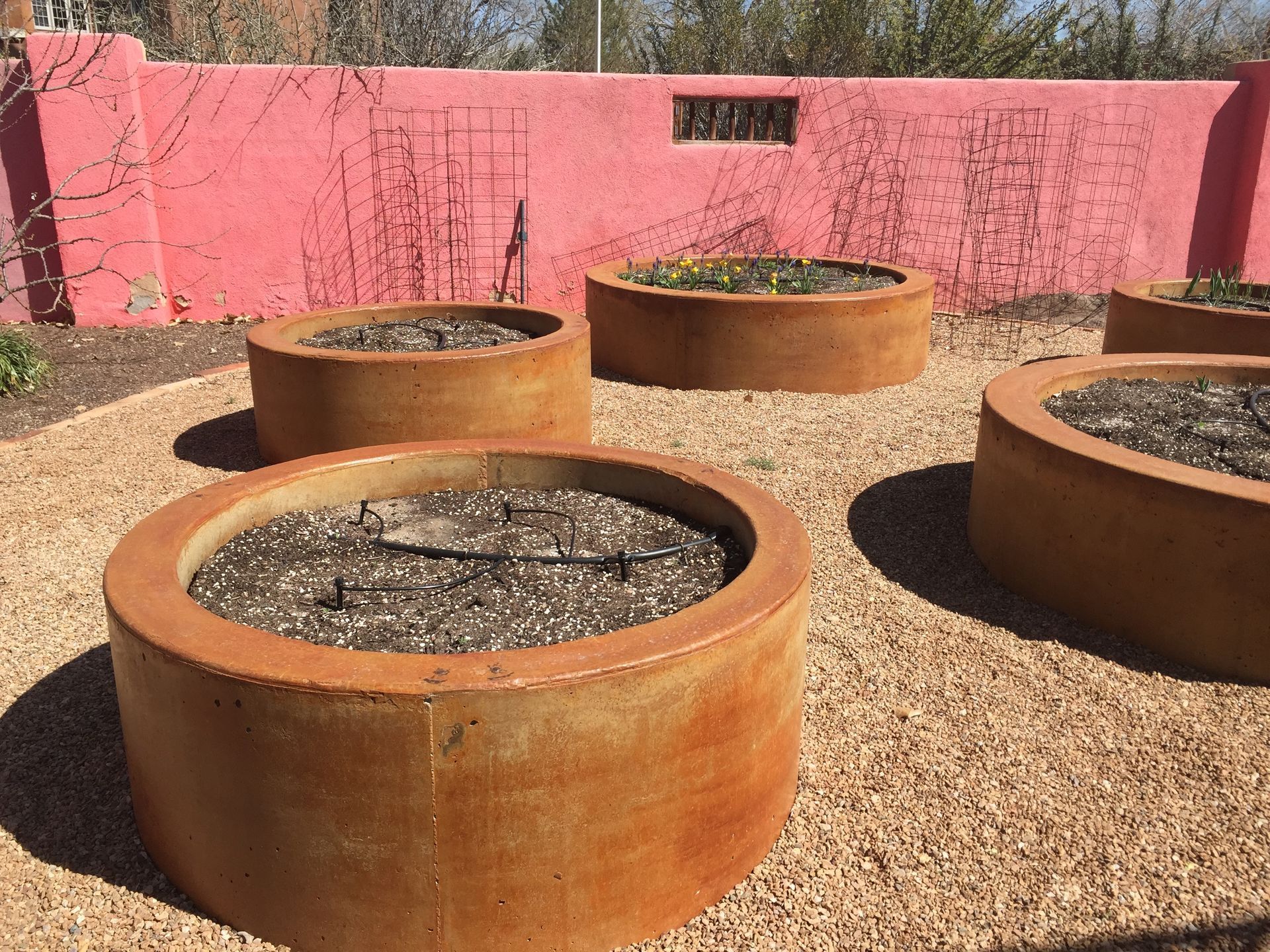Round, rusted planters with dark soil on a bed of gravel in front of a pink wall.