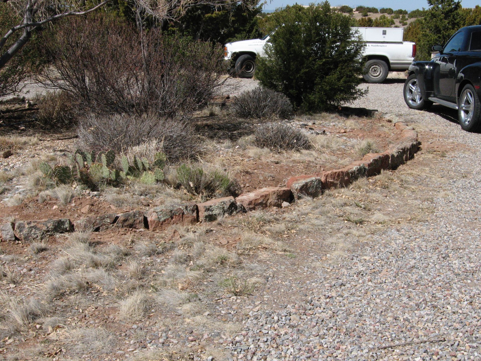 A low, curved brick retaining wall borders dry scrub and gravel driveway; two pickup trucks are parked nearby.