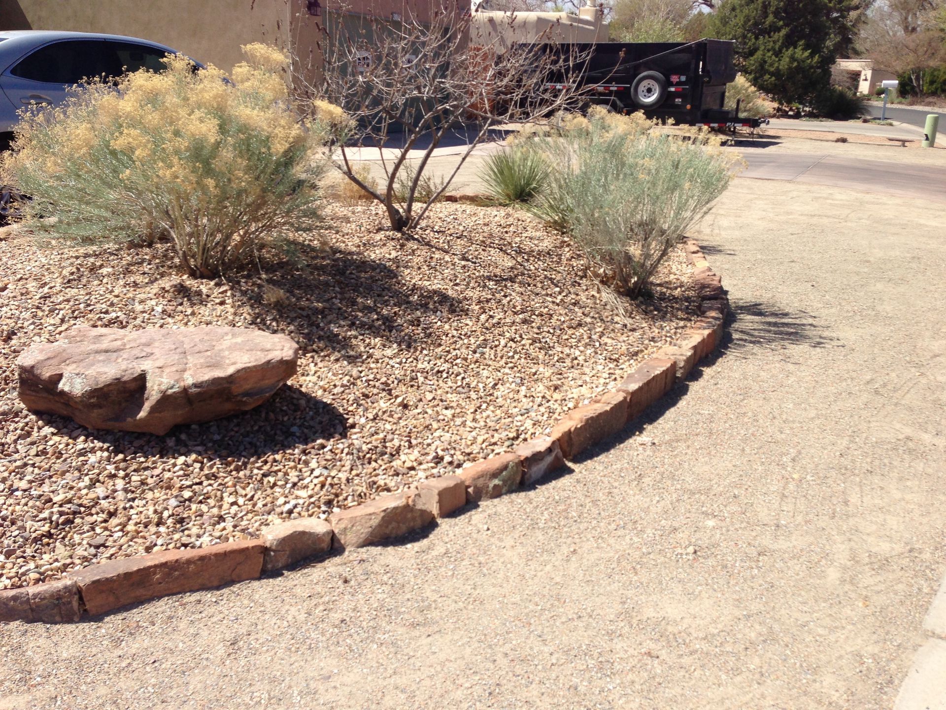 Rock-bordered flower bed with gravel, shrubs, and a large rock, next to a gravel path and driveway.
