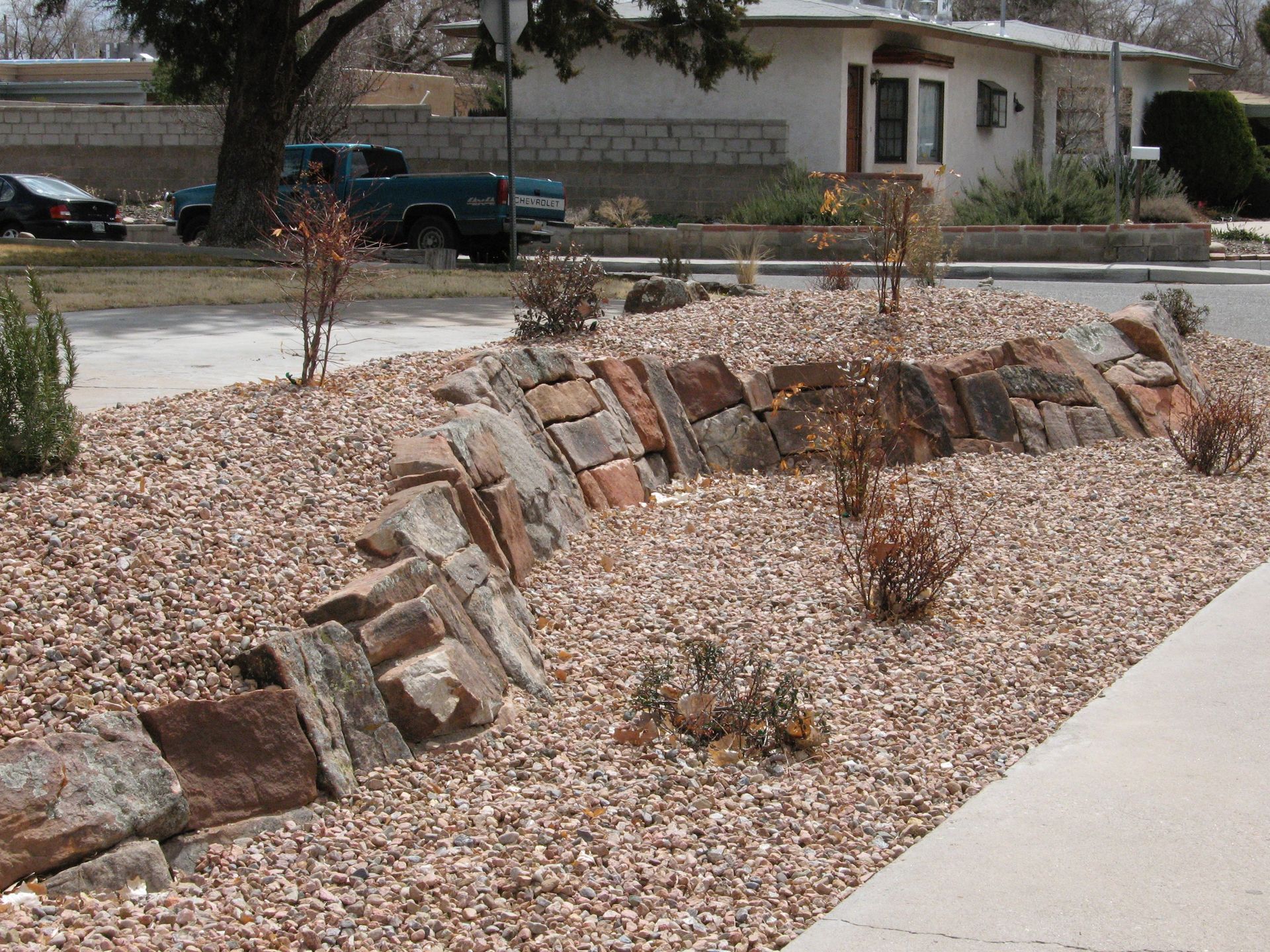 A rock garden with reddish-brown stones, shrubs, and gravel in front of a house on a street.