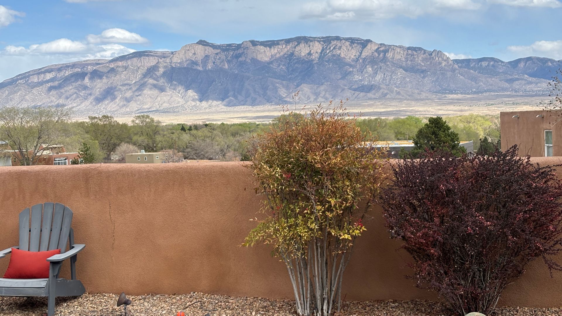 Mountains visible beyond a brown wall, chair, and bushes. Sunny day.