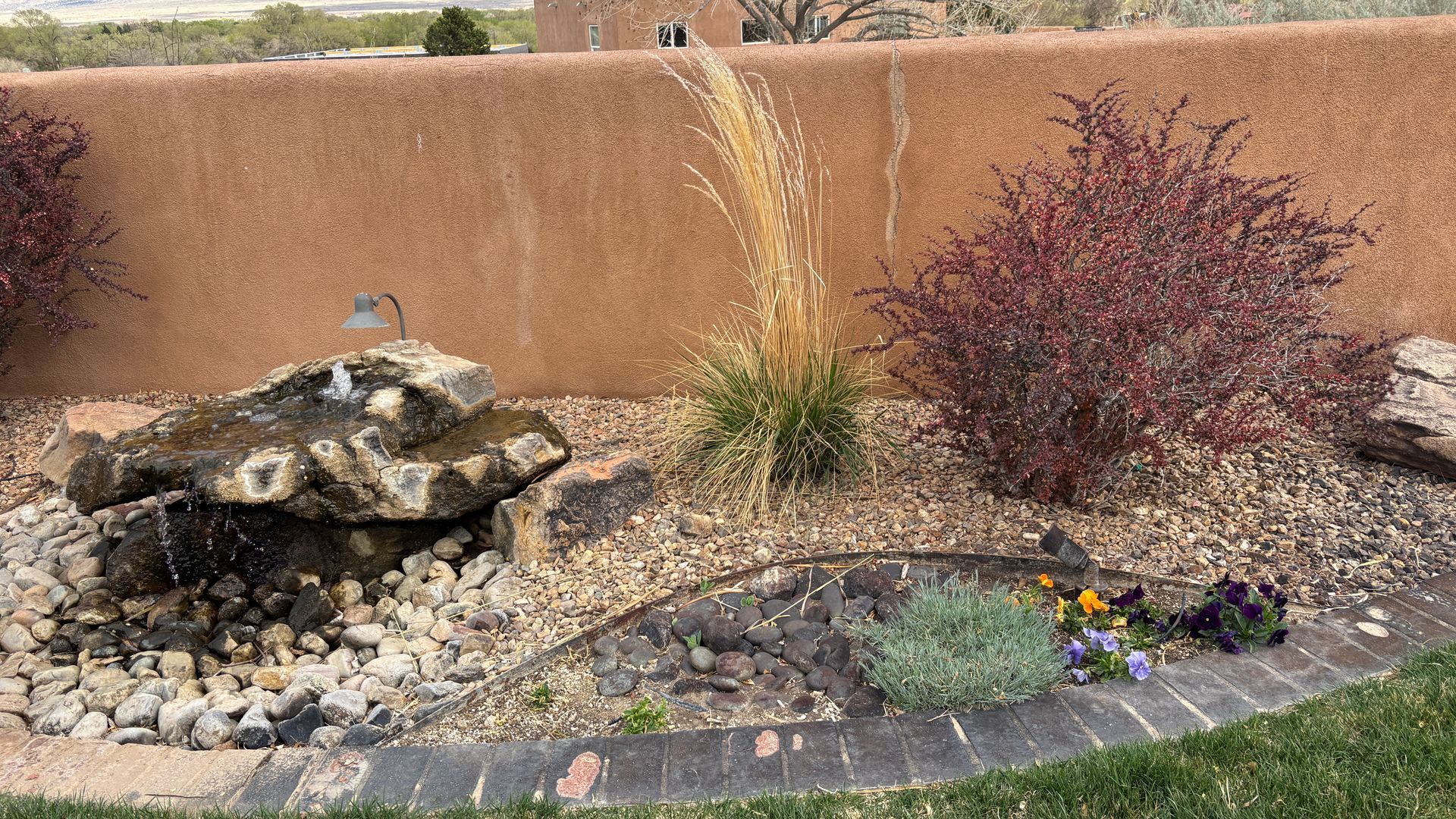 Water fountain with rock formation, landscaping with red and green plants, against a stucco wall.