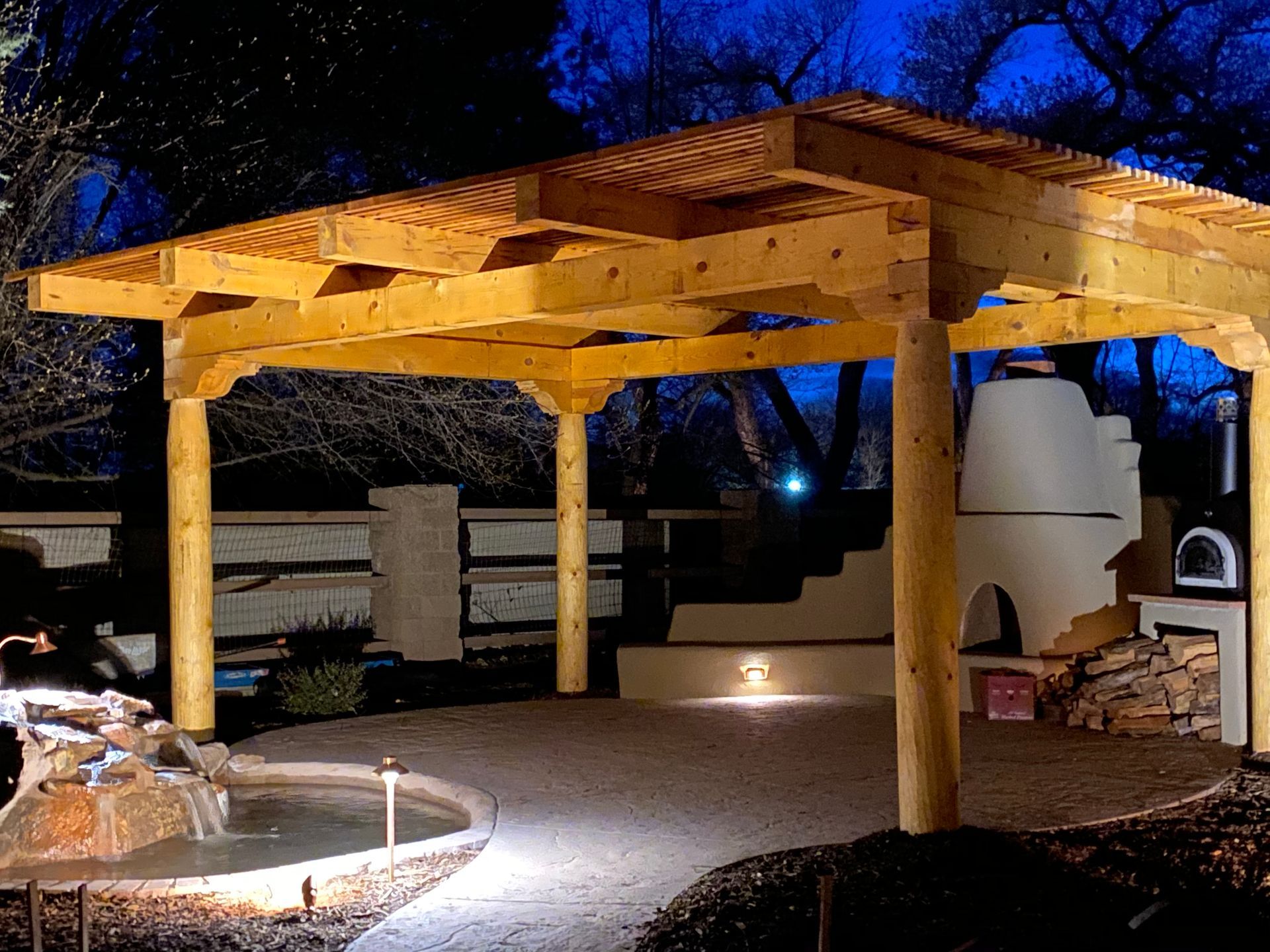Wooden pergola over a patio with a fountain and pizza oven at night.