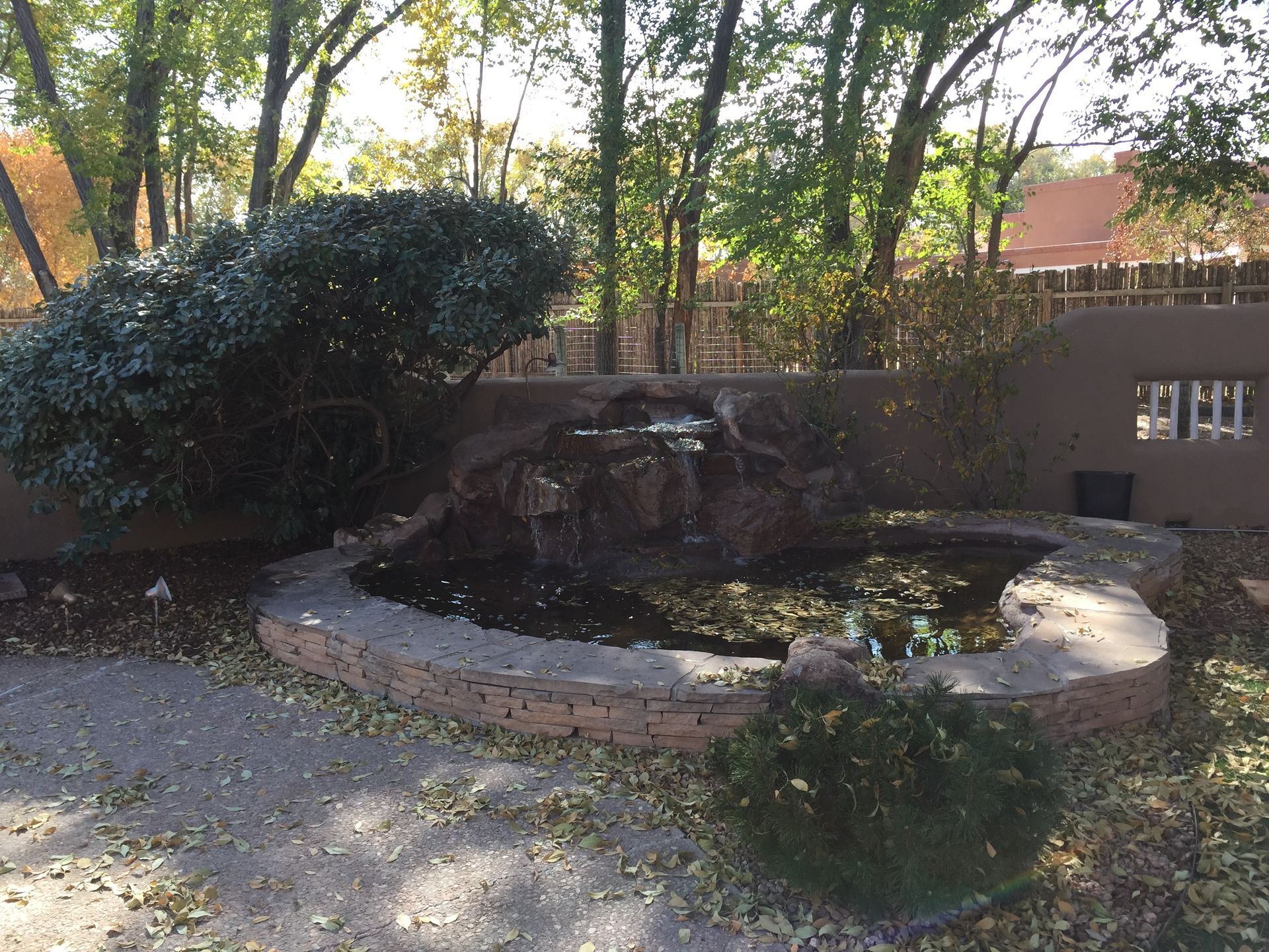 Water feature in a garden setting; a waterfall over rocks into a circular pond, beige wall background, trees.