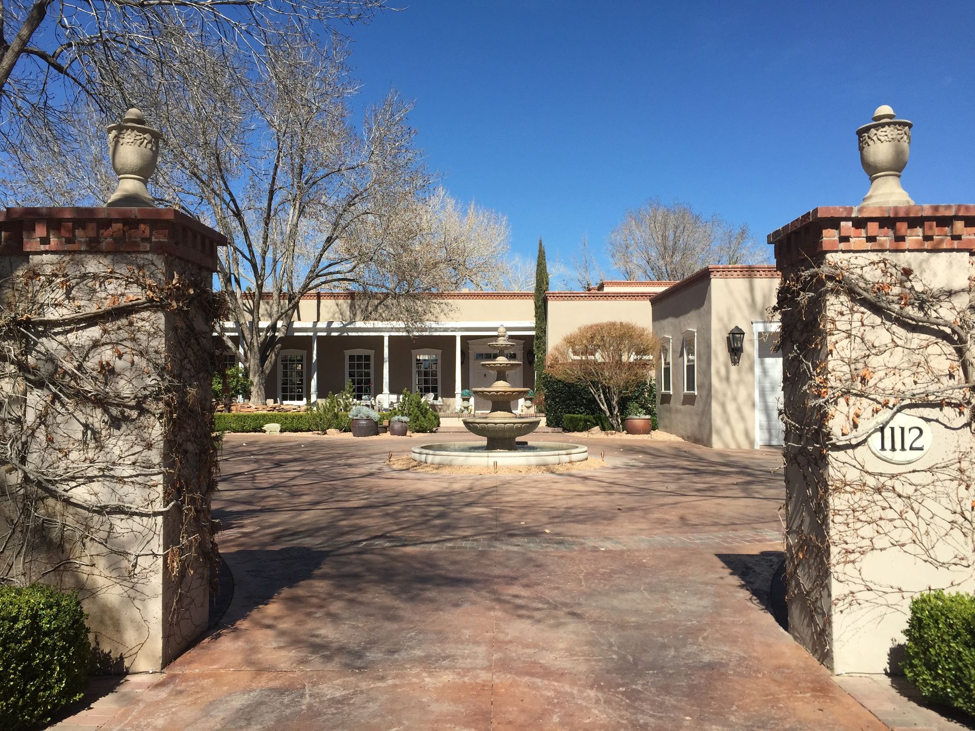 Entryway of a Southwestern-style home, red brick pillars with vines flank a stone driveway, a fountain in the middle.