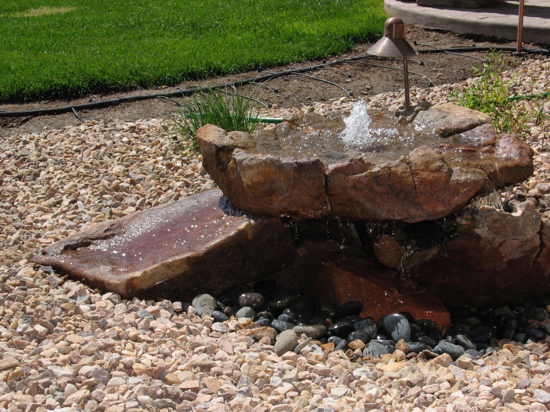 Stone water fountain in a gravel yard, with water spraying into a shallow basin.