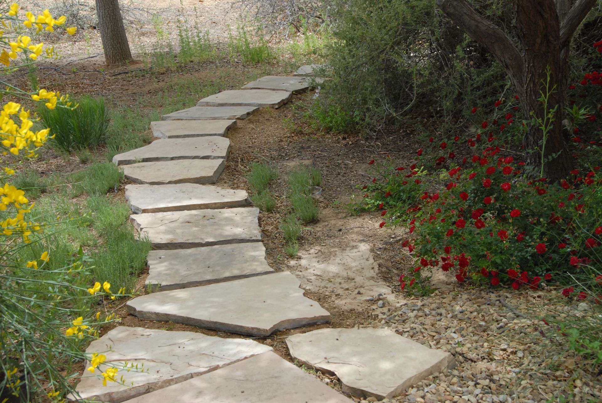 Stone path winding through a garden with yellow and red flowers, surrounded by greenery.