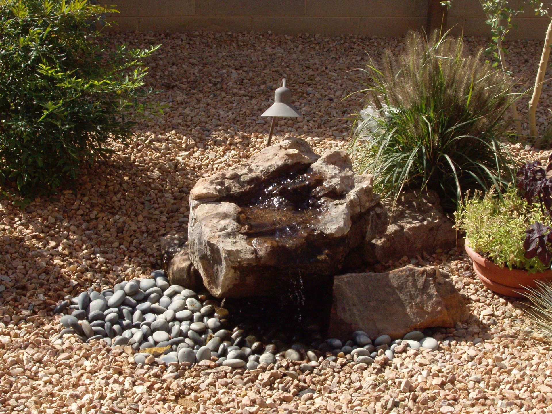 A small rock water fountain in a gravel yard with greenery and a small light.
