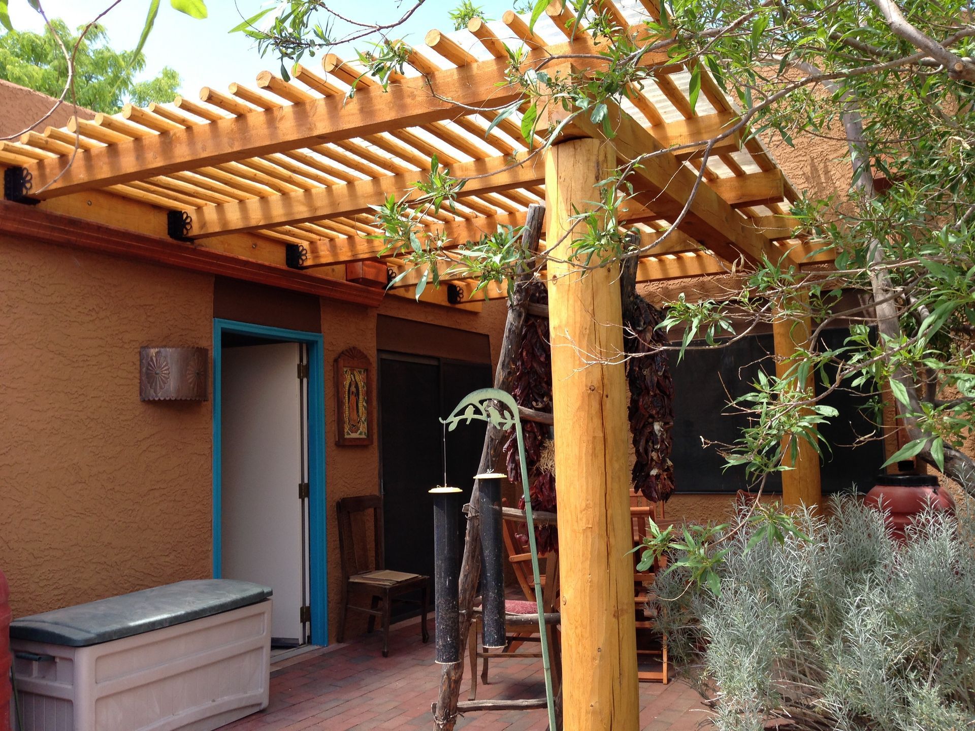 Wooden pergola over a patio; tan stucco building with blue door; tree branches.