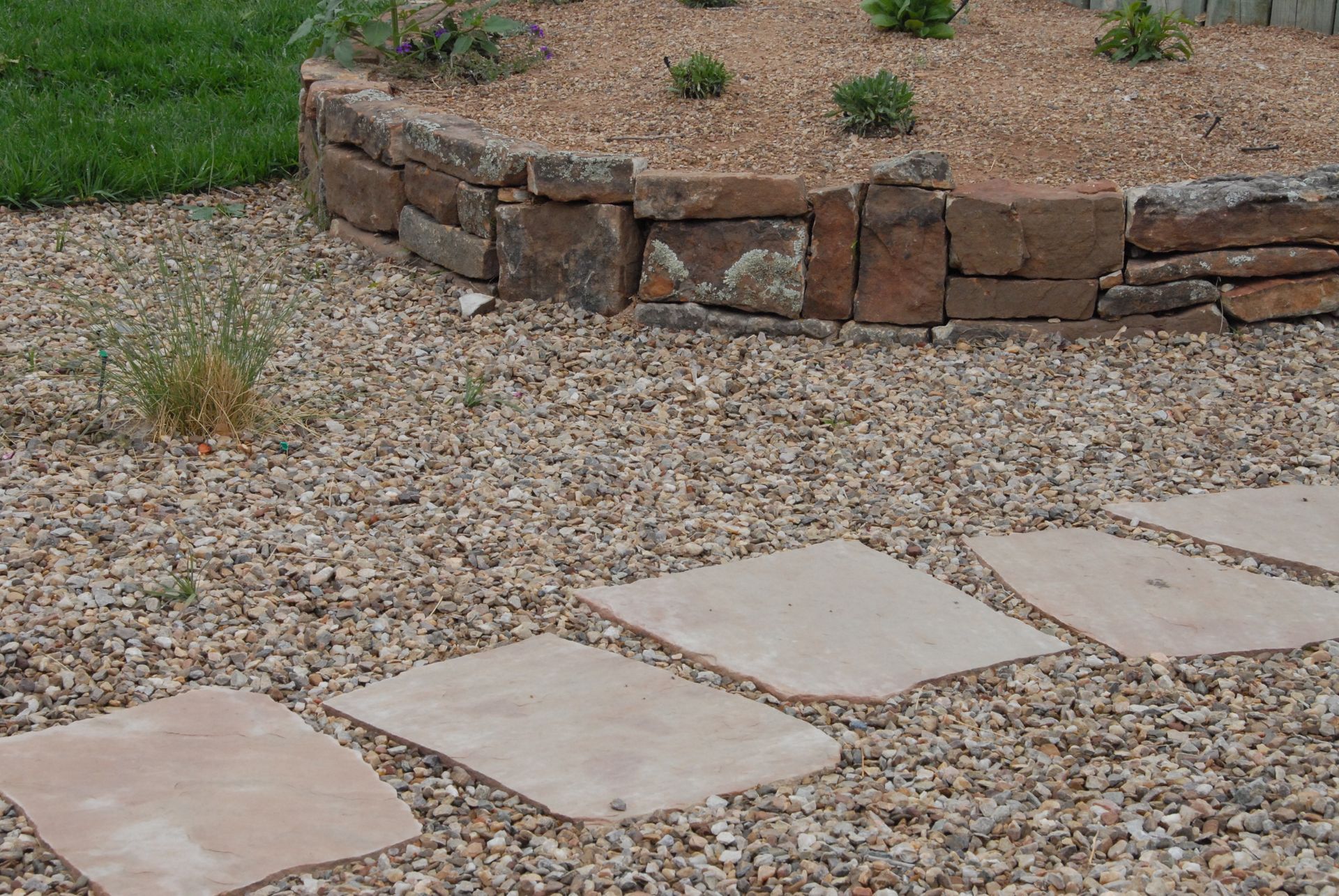 Stone path through gravel leads to a raised garden bed with stone retaining wall.