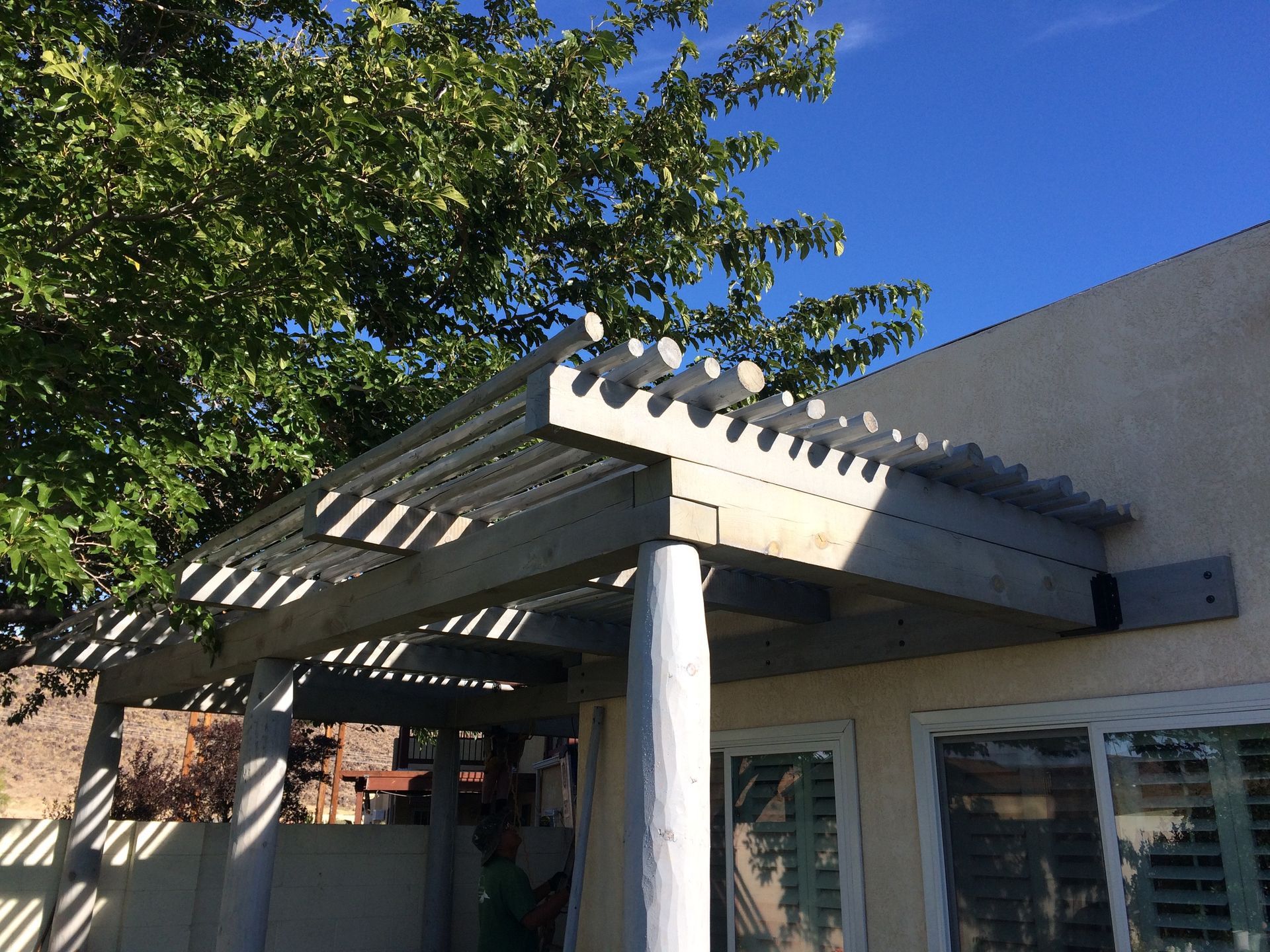 Gray pergola attached to a beige house, with vertical beams and overhead slats, under a sunny blue sky.