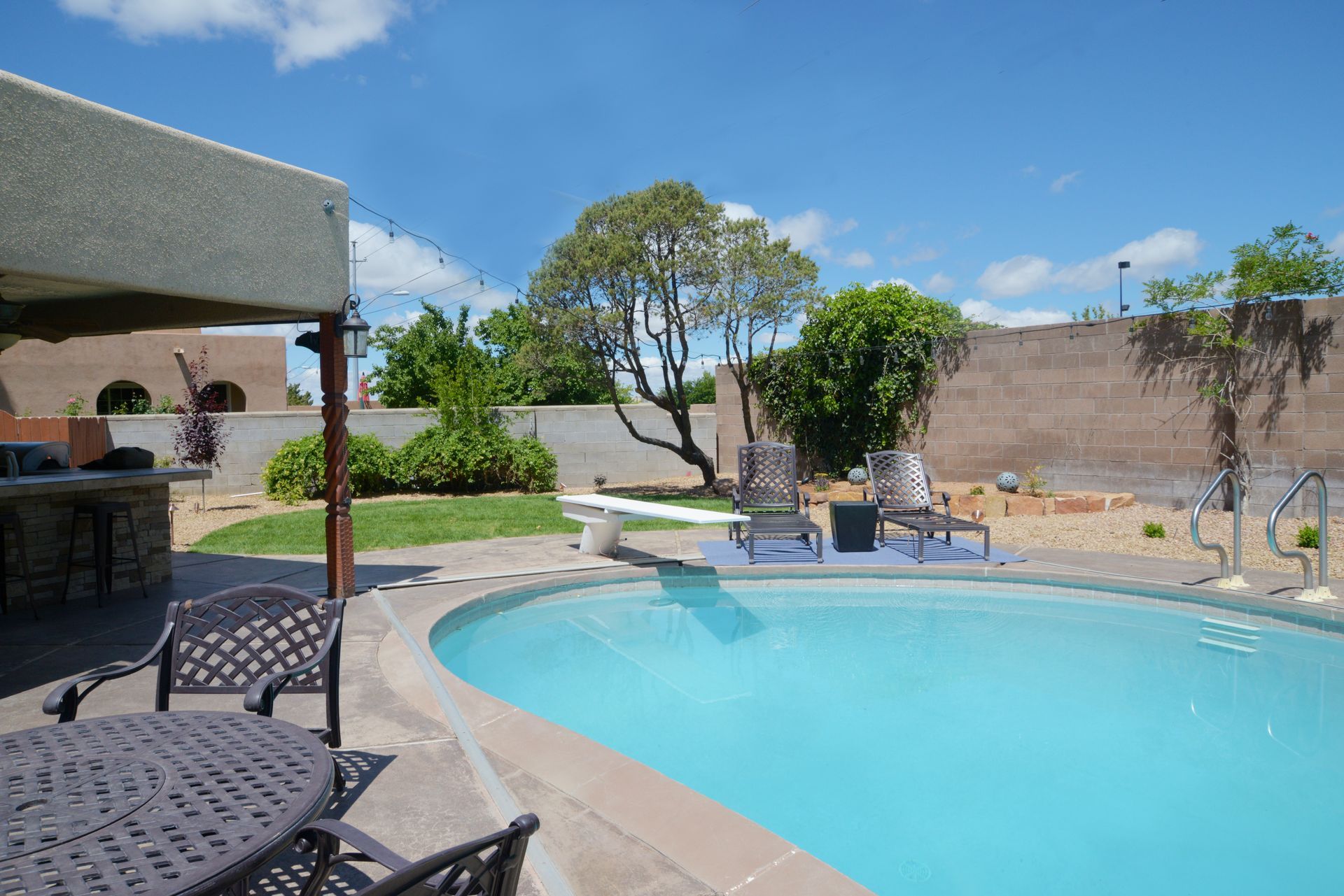 Backyard with a swimming pool, diving board, and patio furniture under a bright blue sky.