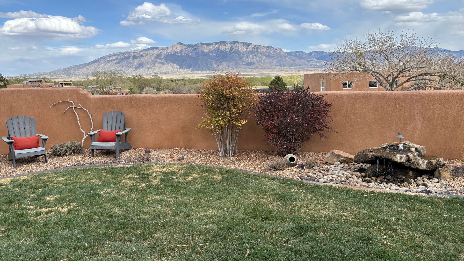 Two Adirondack chairs with red cushions in a grassy backyard with a mountain backdrop.