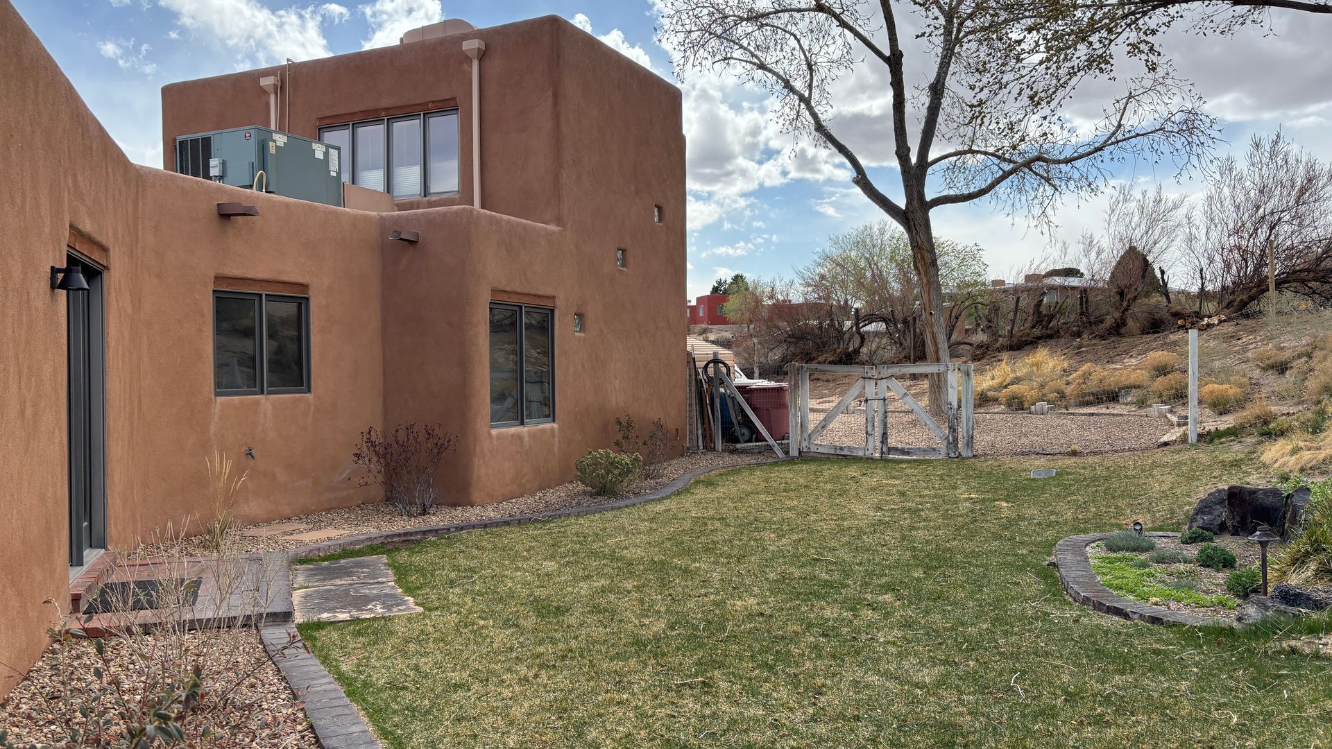 Adobe-style home with a backyard and green lawn, surrounded by a fence and arid landscape under a sunny sky.