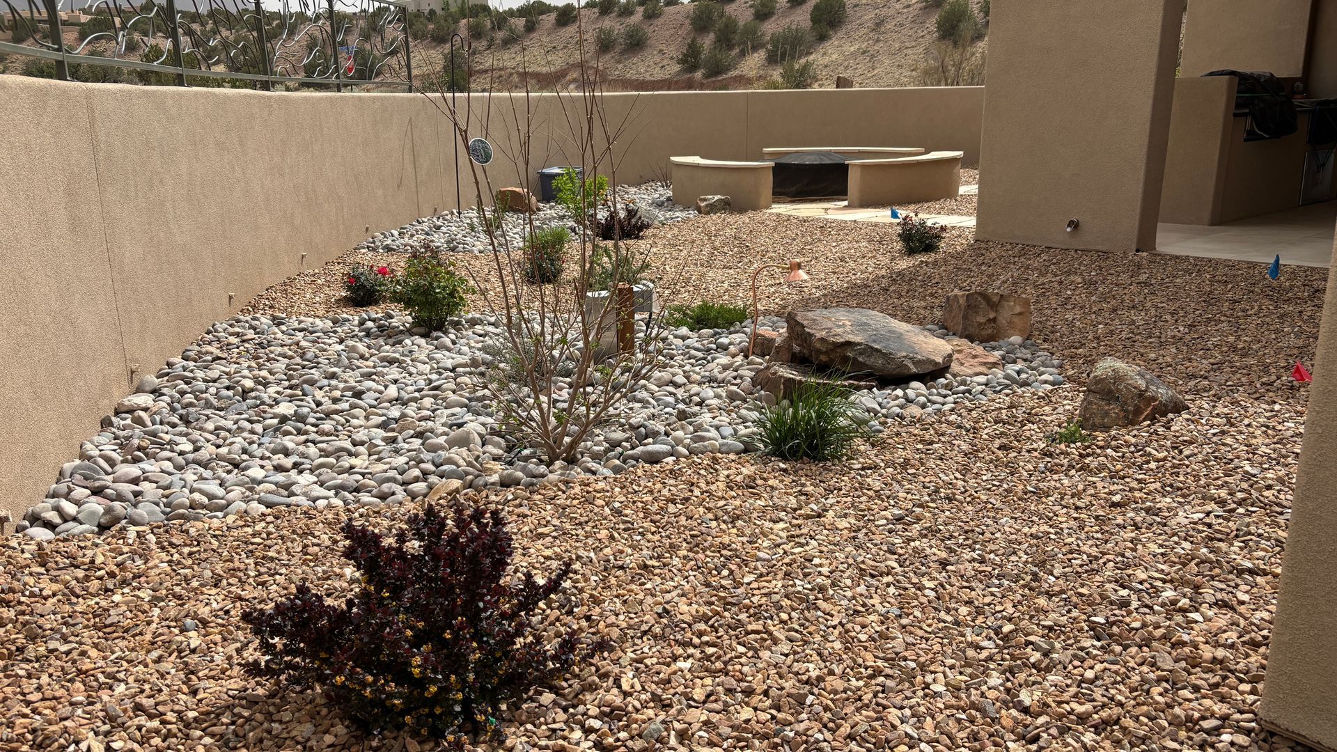 A rock garden with various plants, a fire pit area, and beige walls under a sunny sky.