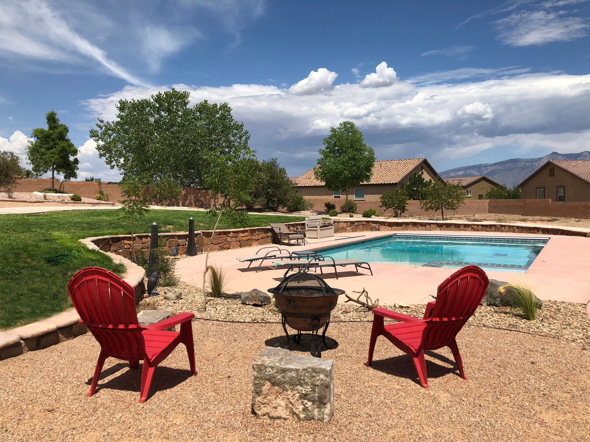 Two red chairs face a fire pit, pool, and houses under a blue sky.