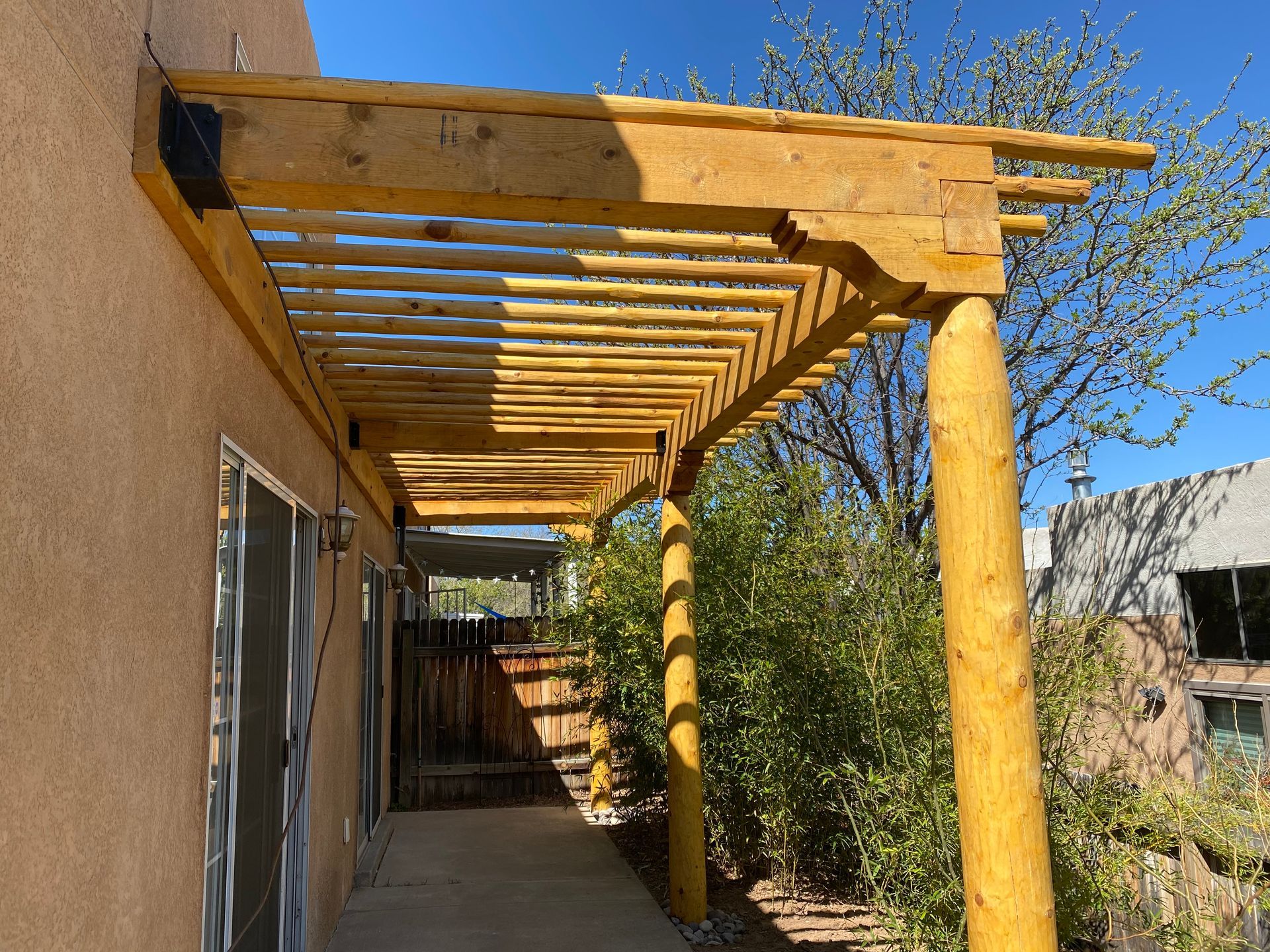 Wooden pergola extending from a tan stucco wall, supported by round posts. Sunny outdoor setting.