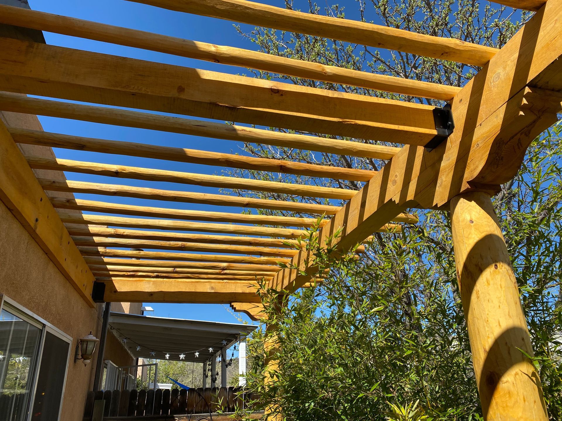 Wooden pergola with horizontal beams against a clear blue sky, attached to a tan building with a patio.