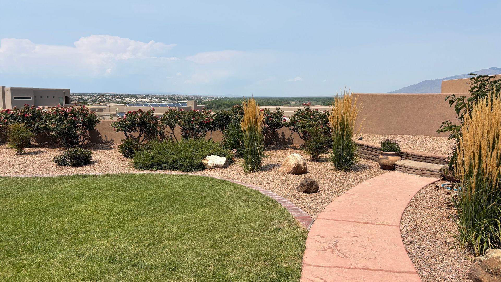 A landscaped backyard with a green lawn, stone path, and gravel beds, overlooking a distant cityscape and mountains under a blue sky.