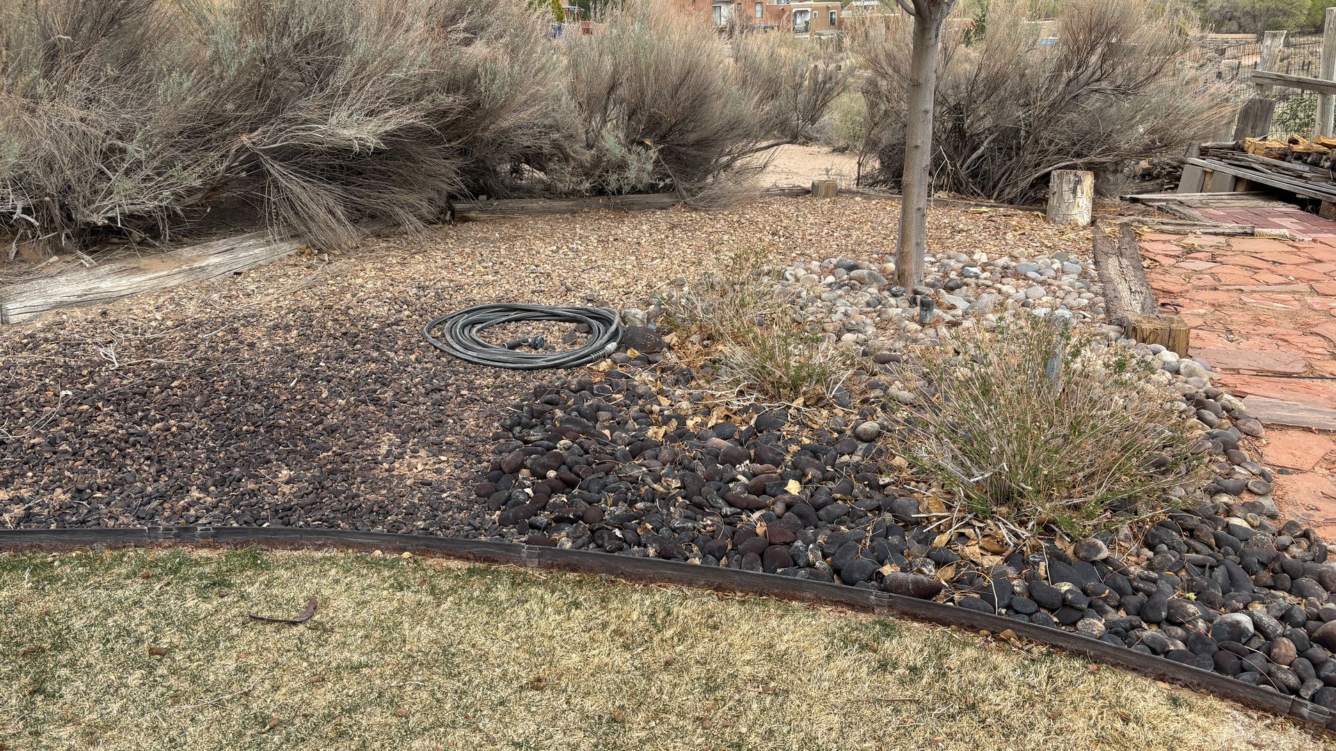 A rock garden with dry brush, gravel, and a dark border next to a lawn and brick patio.
