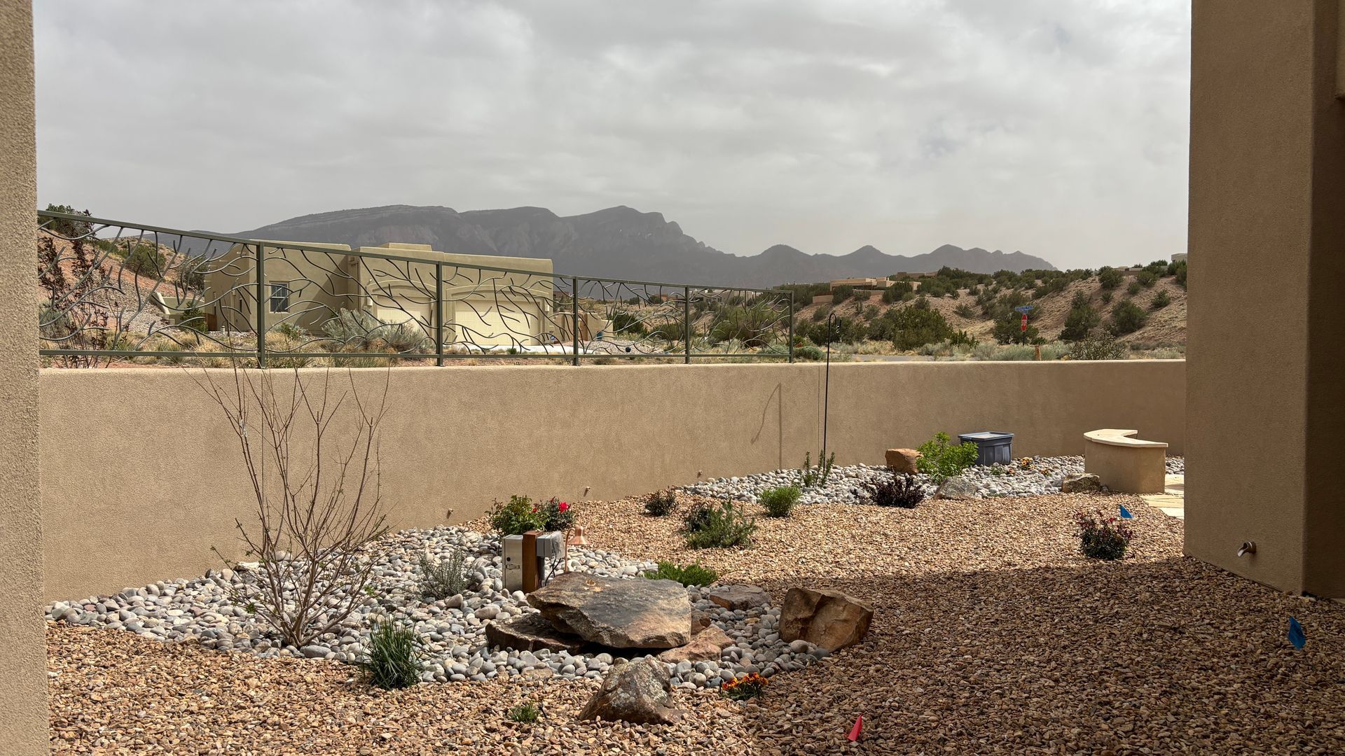 Backyard with desert landscaping, tan wall, and mountains in the background under a cloudy sky.
