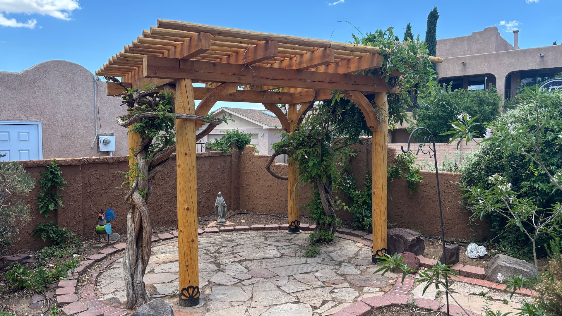 Wooden pergola in a garden, with stone patio, surrounded by greenery and a stucco wall.