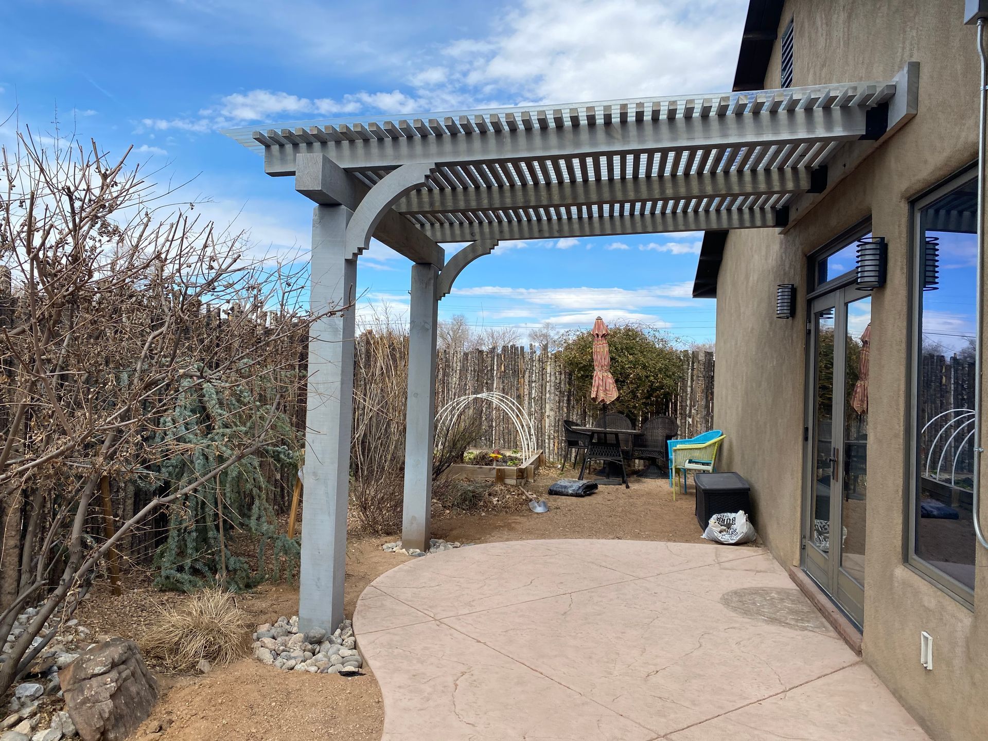 Pergola extends from a beige house over a curved patio, with a fence and bushes in the background.