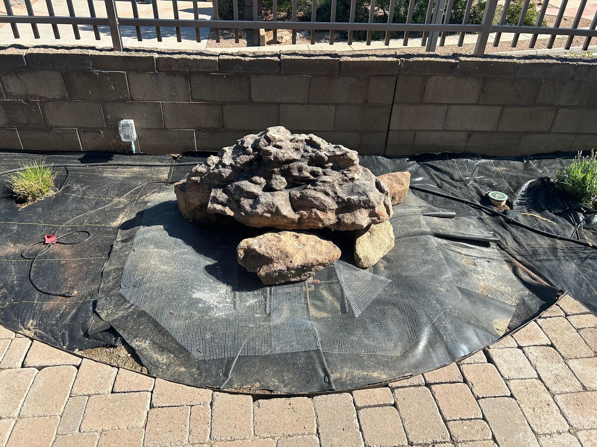 Large rocks on black landscaping fabric, set in a brick-edged garden bed next to a fence and a brick wall.