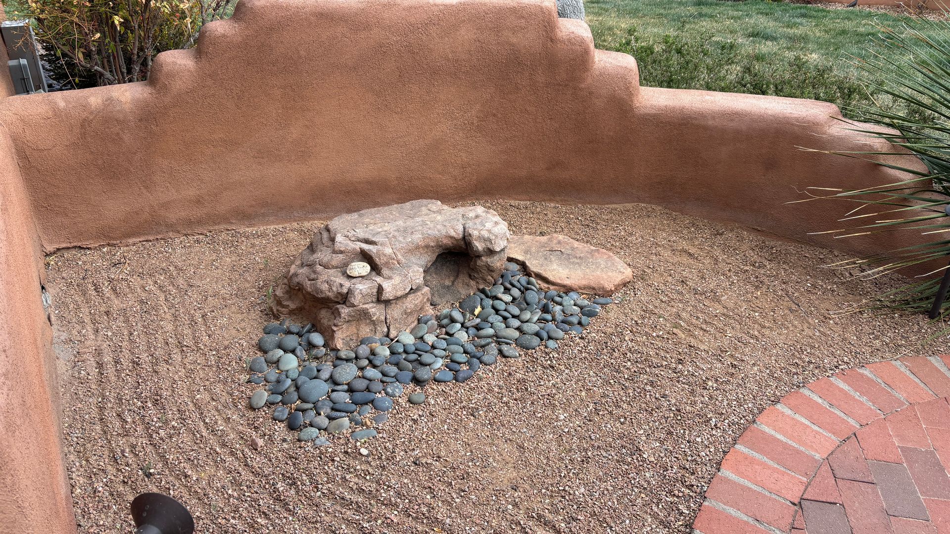 Rock formation in a gravel bed within a walled area, featuring a brick patio and tan adobe walls.