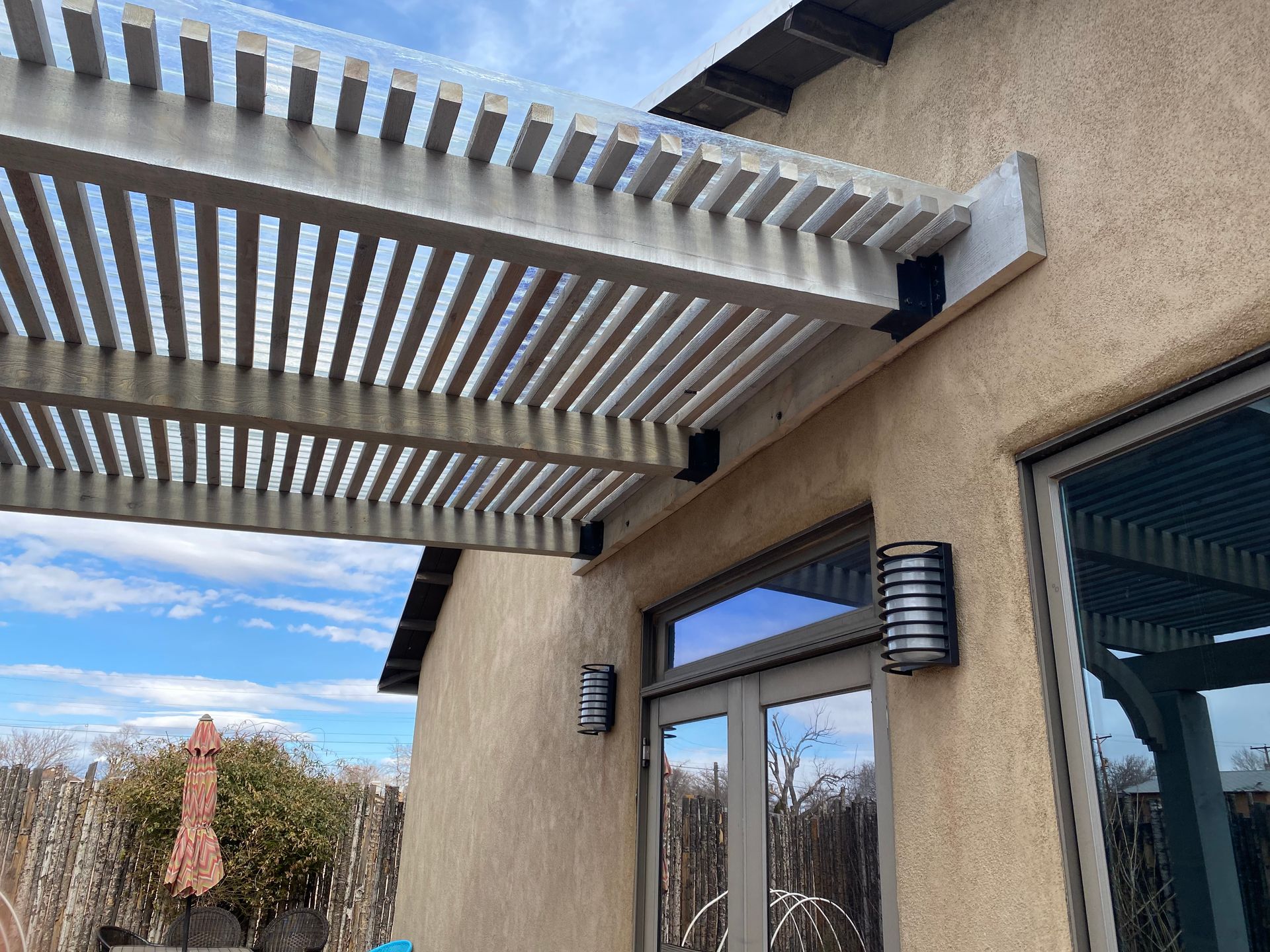 Pergola attached to stucco house with glass doors and outdoor sconces.