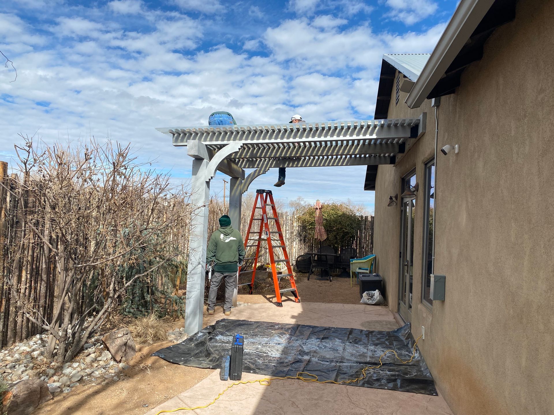 Person installing a pergola on a patio next to a tan house under a partly cloudy sky.