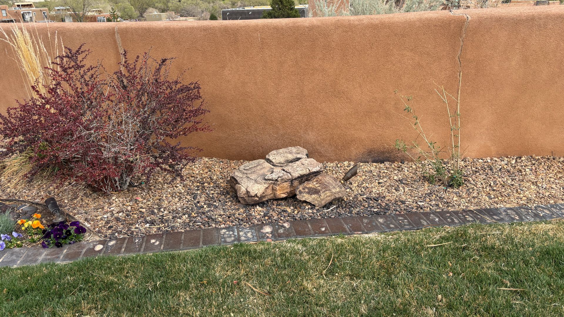 A grassy lawn leads to a low rock-lined planting bed with bushes and a reddish-brown stucco wall in the background.