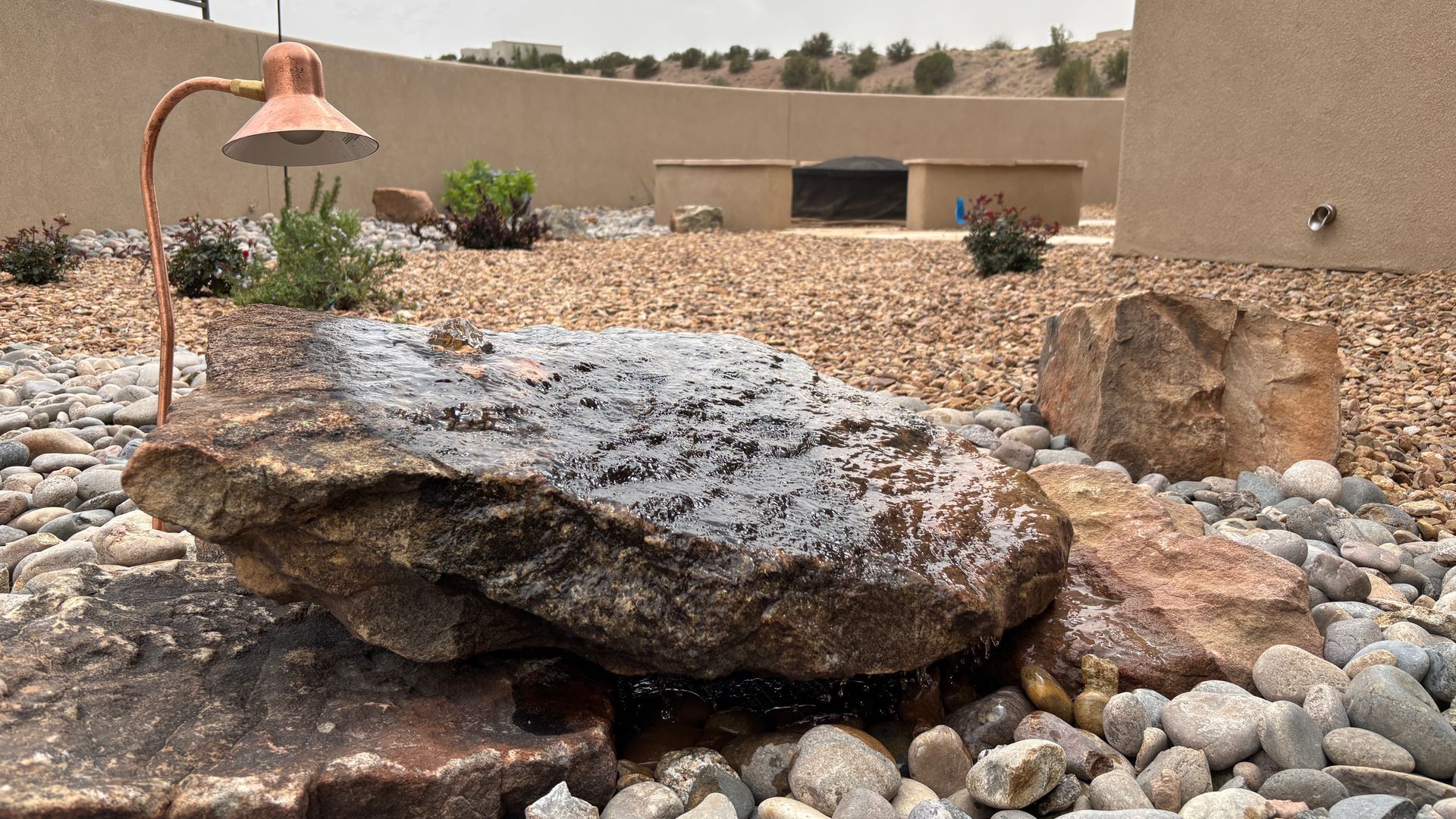 A rock water fountain in a yard with brown pebbles, a copper lamp, and a backdrop of a wall and hills.