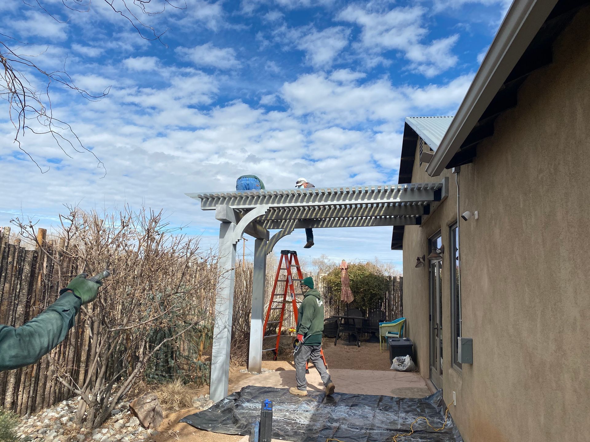 Man working on a white pergola attached to a tan building. Ladder, tools, and cloudy sky visible.