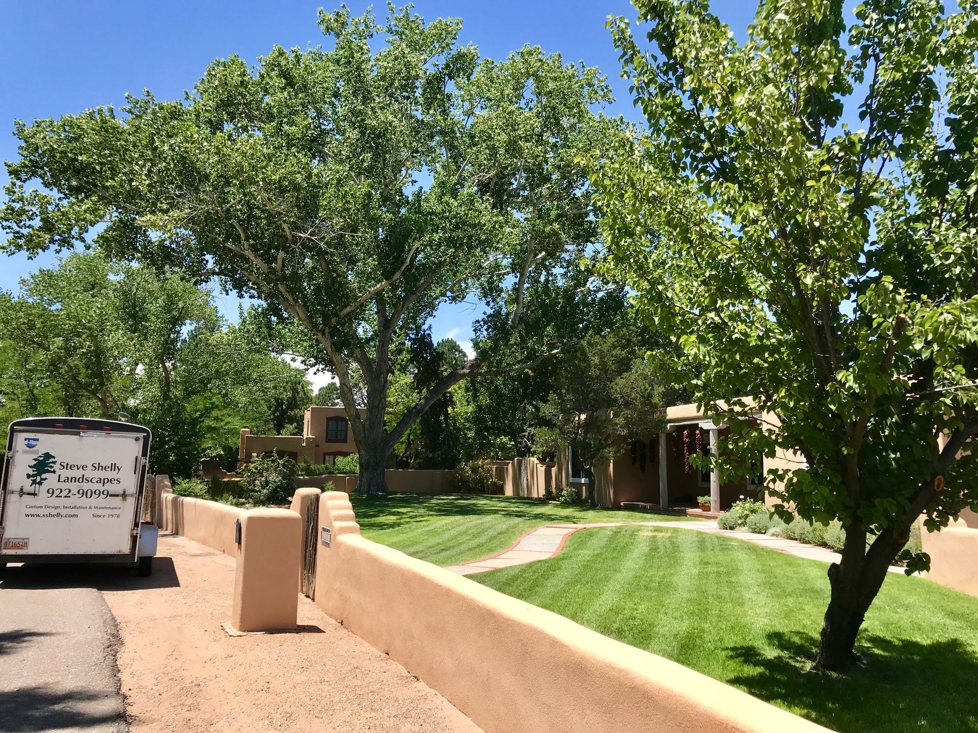 Lawn care truck parked on a street with stucco walls, grass, and trees under a blue sky.